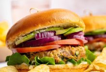 A chicken burger served on a brioche bun topped with lettuce, tomato, red onion and avocado. The burger is served with a side of potato chips.