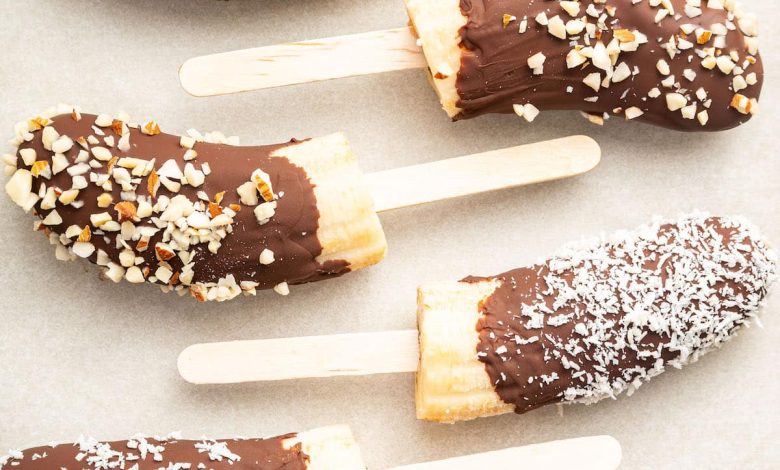 Frozen banana halves on wooden sticks, dipped in chocolate and topped with various toppings including rainbow sprinkles, chopped nuts, and shredded coconut.