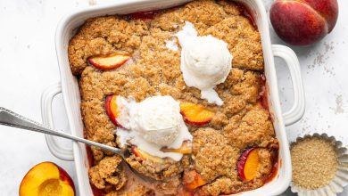 Freshly baked healthy peach cobbler in a white baking dish topped with vanilla ice cream and a spoon scooping out a serving.