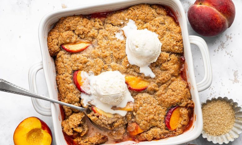 Freshly baked healthy peach cobbler in a white baking dish topped with vanilla ice cream and a spoon scooping out a serving.