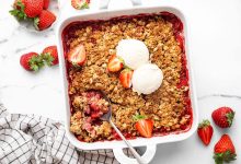 Baked strawberry crisp topped with two scoops of vanilla ice cream and fresh strawberry halves. There is a spoon scooping out some of the crisp.