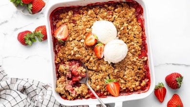 Baked strawberry crisp topped with two scoops of vanilla ice cream and fresh strawberry halves. There is a spoon scooping out some of the crisp.