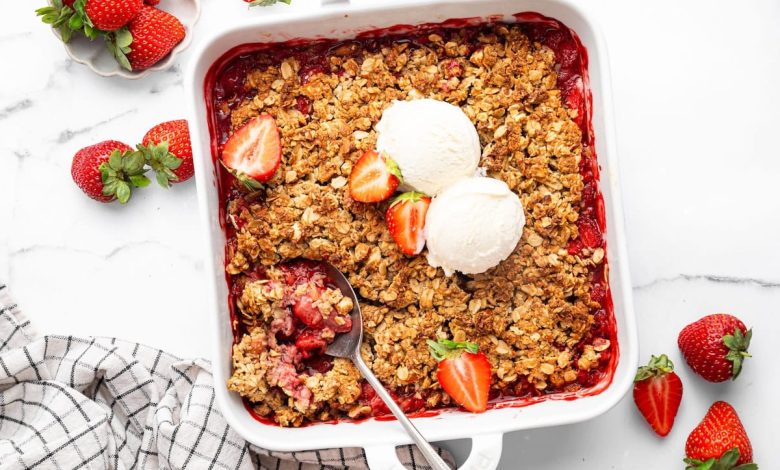 Baked strawberry crisp topped with two scoops of vanilla ice cream and fresh strawberry halves. There is a spoon scooping out some of the crisp.